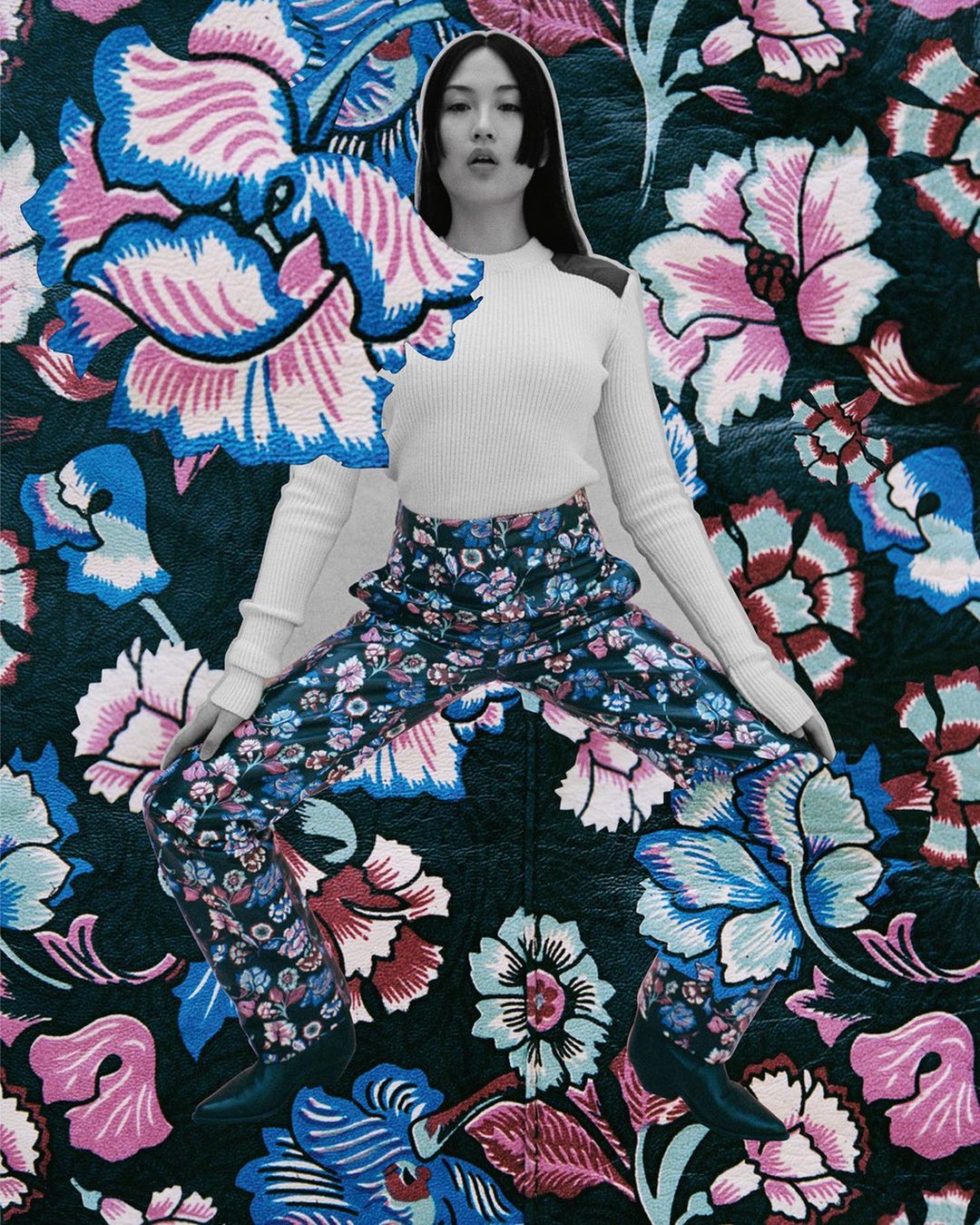 Woman in a white long-sleeved shirt and blue floral pants strikes a pose against a backdrop of large floral patterns in blues and pinks.