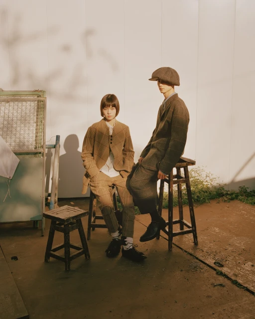 Two fashion models, a man and a woman, pose while wearing brown tweed suits and sitting on wooden stools in a photo studio.