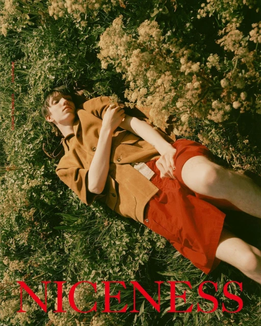 Young man lies in sunlit field of flowers wearing caramel jacket and red shorts in relaxed pose, captured with eye-level angle.