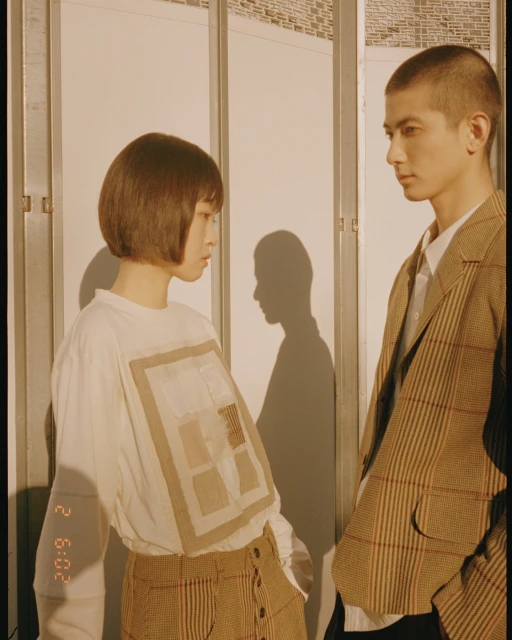 Indoor portrait of an Asian man and woman in fashionable neutral-toned outfits standing close, with graphic tee and shadow.