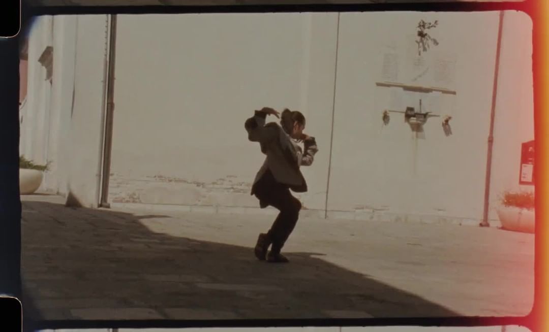 Adult in business attire dancing outdoors, framed by buildings and open sky, capturing dynamic movement and urban setting.