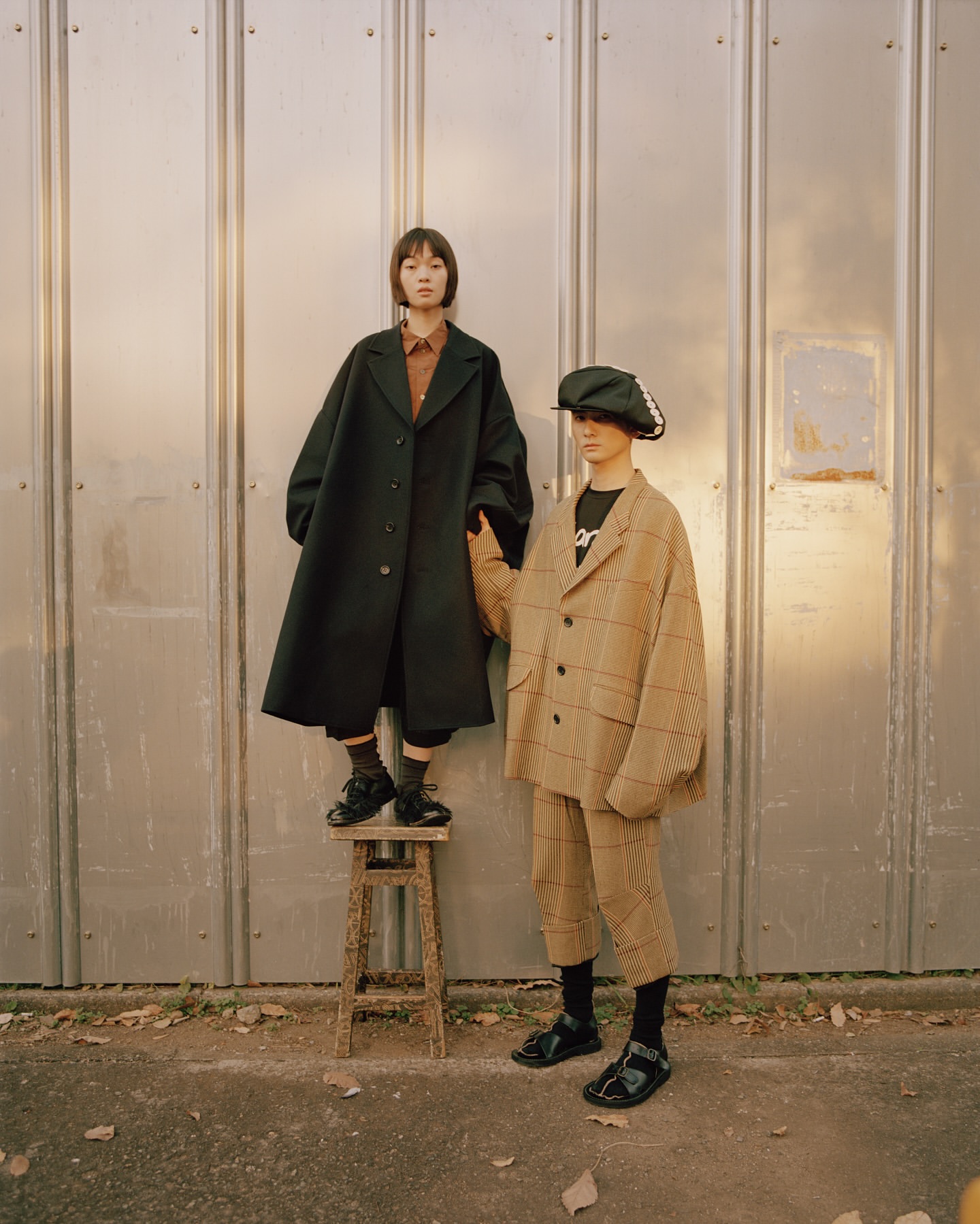 Two women stand outdoors in oversized coats, one atop a wooden stool against a metal wall, showcasing fall fashion apparel.