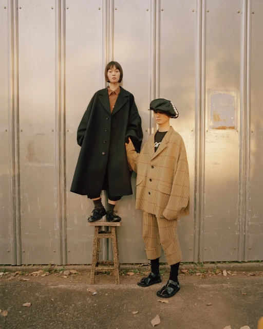 Two women stand outdoors in oversized coats, one atop a wooden stool against a metal wall, showcasing fall fashion apparel.