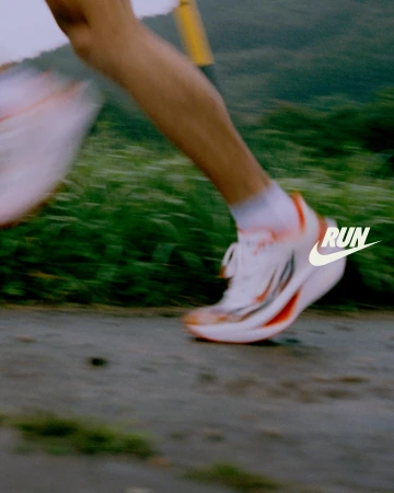 Person running on a path wearing white and orange running shoes, showing motion blur, with grass and greenery in the background.
