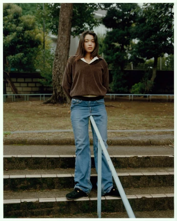 Young woman in brown sweatshirt and blue jeans standing outdoors on stone steps by a metal railing, photographed by Piczo.