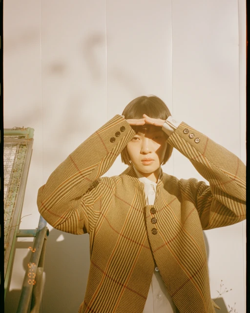 Adult female in a brown plaid jacket poses with hands above head, set against a plain wall, with a ladder and vintage prop.