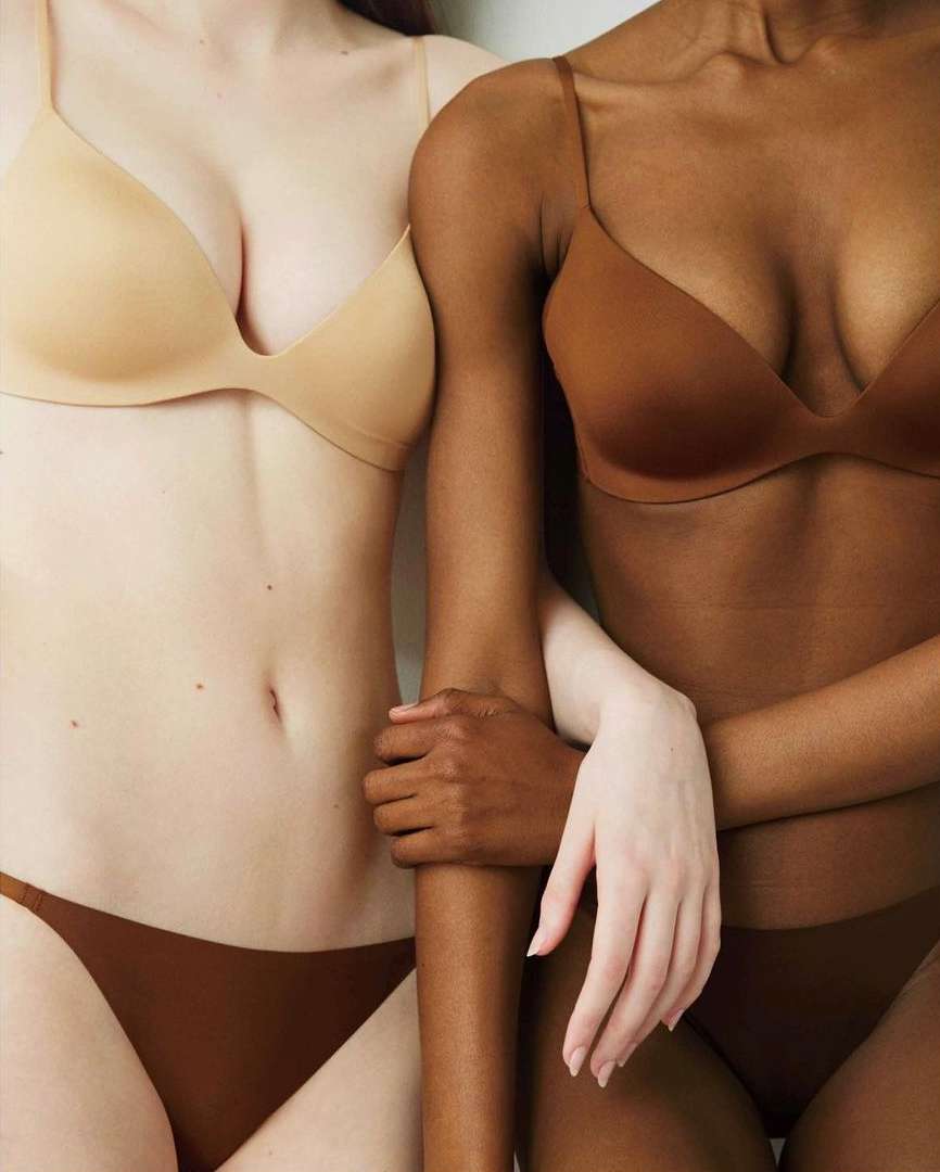 Two women of different ethnicities pose close together in matching bras and underwear, showcasing diverse skin tones in a studio setting.