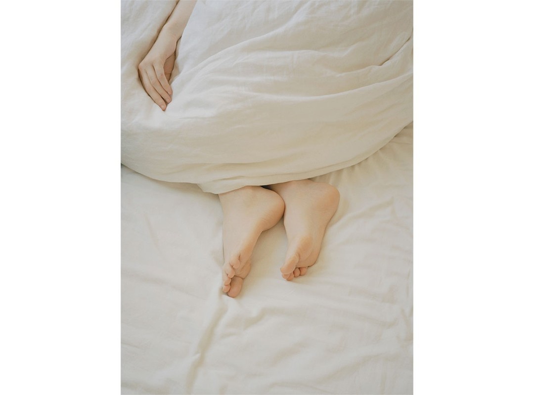 Close-up of Caucasian woman's feet and hand protruding from under a white duvet on a white bed in an indoor setting.