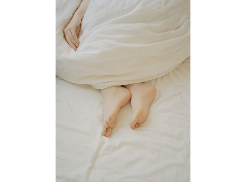 Close-up of Caucasian woman's feet and hand protruding from under a white duvet on a white bed in an indoor setting.