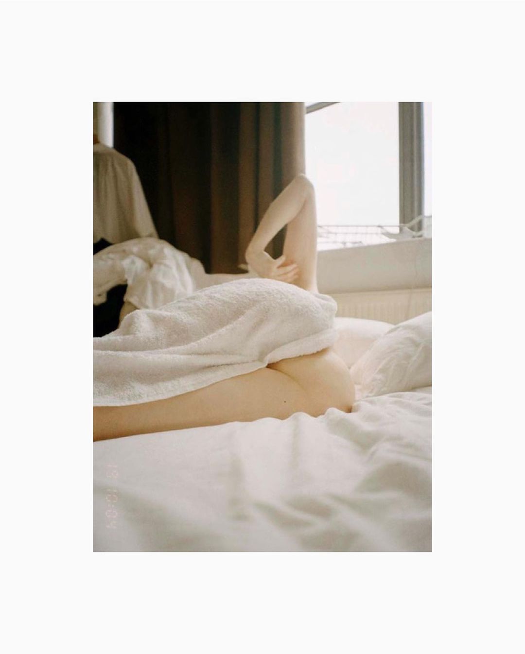 Person lies in bed covered with a white towel next to a window with sheer curtains and dark brown curtains with clothing rack behind.