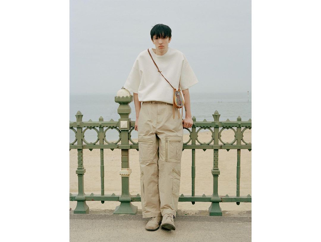 Male model wearing a white t-shirt and beige cargo pants with a shoulder bag, posing in front of a green metal fence and beach.