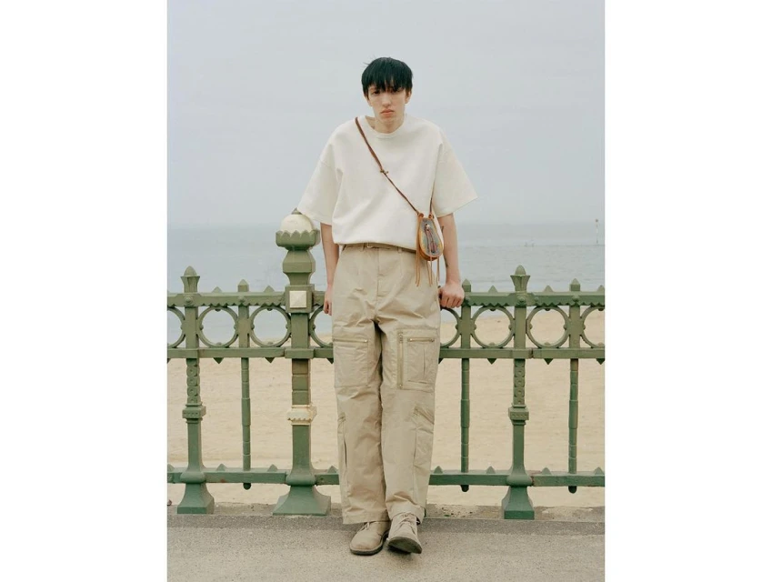 Male model wearing a white t-shirt and beige cargo pants with a shoulder bag, posing in front of a green metal fence and beach.