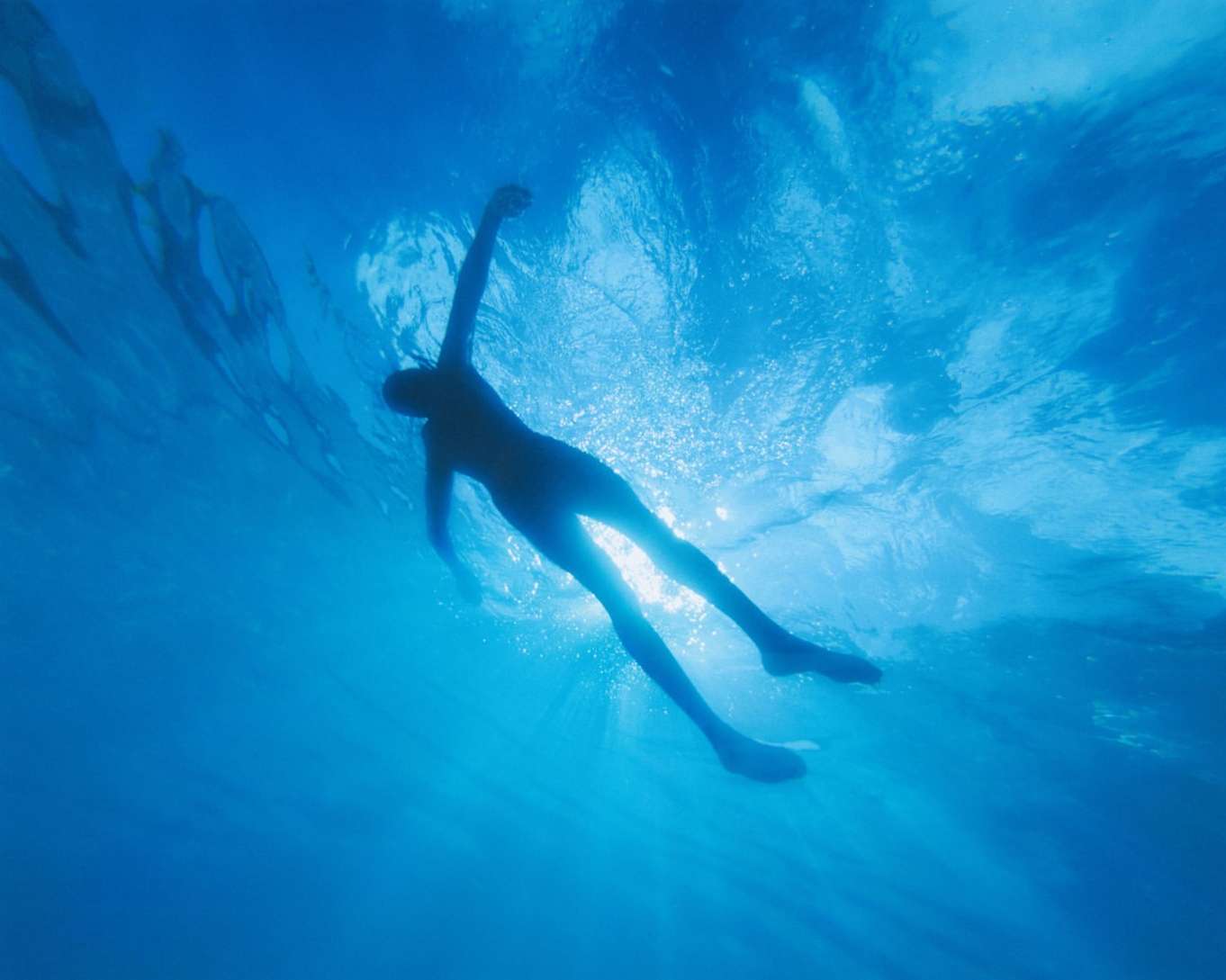 Underwater shot featuring a lone adult female swimmer in silhouette, surrounded by a vibrant blue ocean and sunlight filtering through the surface.