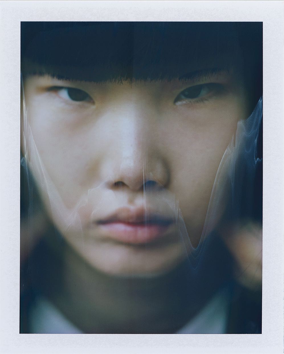Close-up portrait of a young Asian individual wearing a black hat, framed by a hazy, dreamlike effect on a polaroid background.