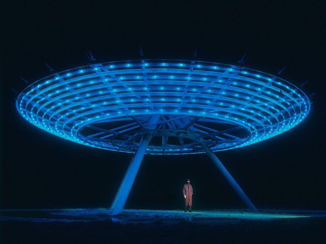 Person in red stands beneath large blue UFO-shaped light sculpture at night, creating a surreal and striking visual contrast.