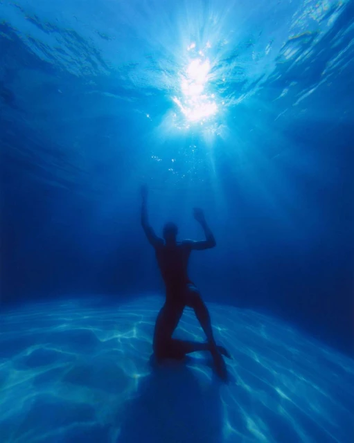 Person underwater in the ocean kneeling on the sand with arms raised towards the sunlight shining through the surface of the water.