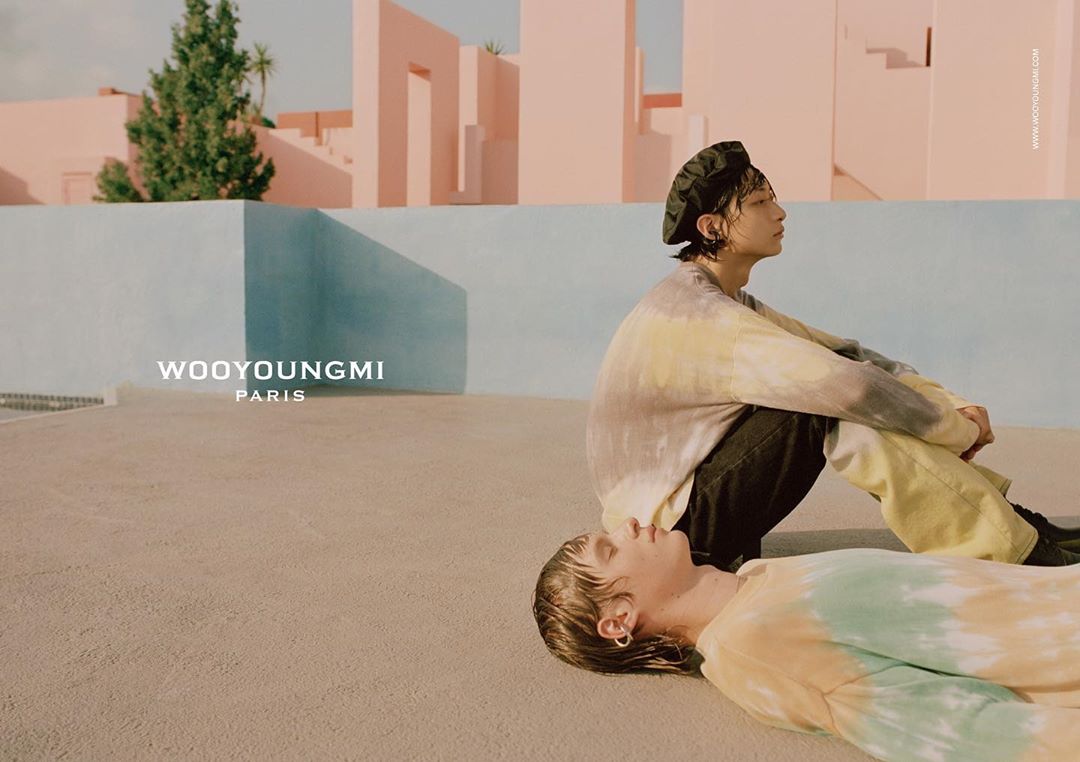 Two young adult models wear tie-dye tops outdoors in a minimalist setting with pink and blue walls, showcasing a contemporary fashion aesthetic.