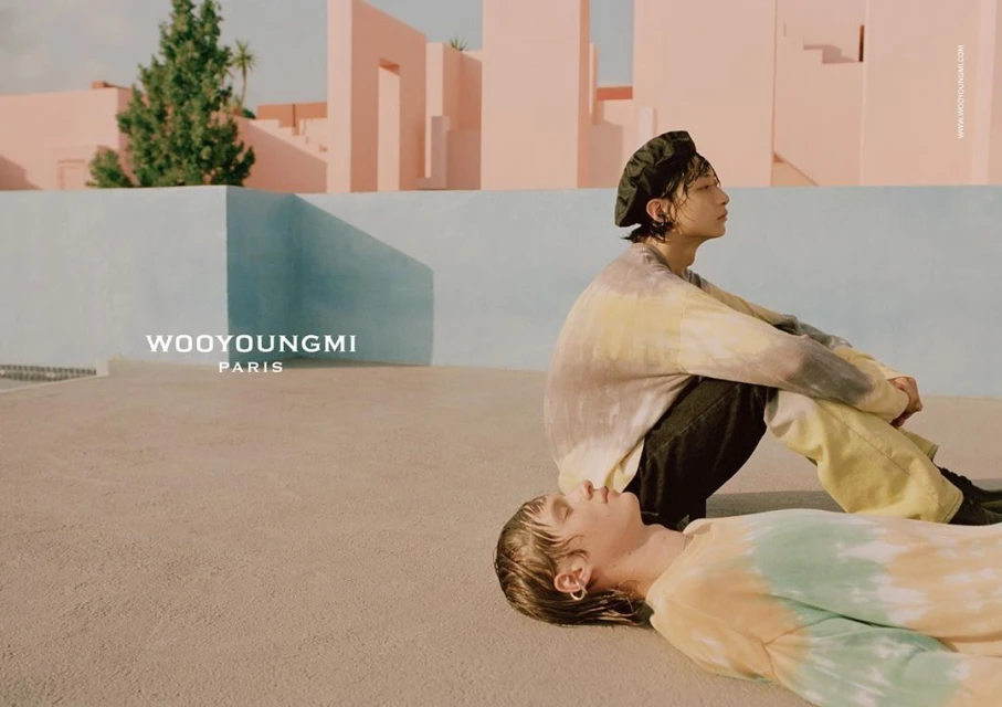 Two young adult models wear tie-dye tops outdoors in a minimalist setting with pink and blue walls, showcasing a contemporary fashion aesthetic.