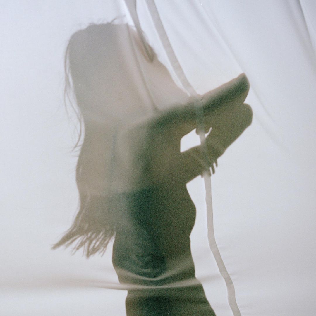 Silhouette of a woman behind a translucent white curtain, hand raised, soft lighting, creating an ethereal and artistic aesthetic.