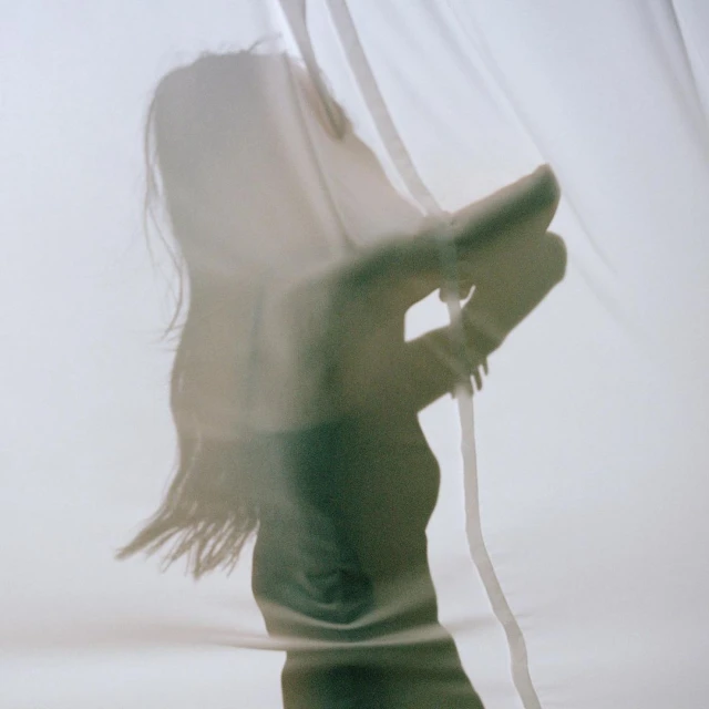 Silhouette of a woman behind a translucent white curtain, hand raised, soft lighting, creating an ethereal and artistic aesthetic.
