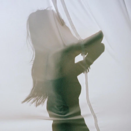Silhouette of a woman behind a translucent white curtain, hand raised, soft lighting, creating an ethereal and artistic aesthetic.