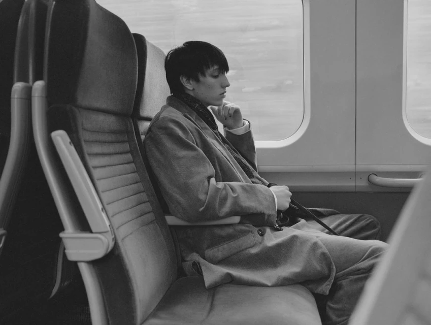Monochrome portrait of a person in a suit seated on a train by a window, with natural lighting emphasizing textures and contemplative mood.