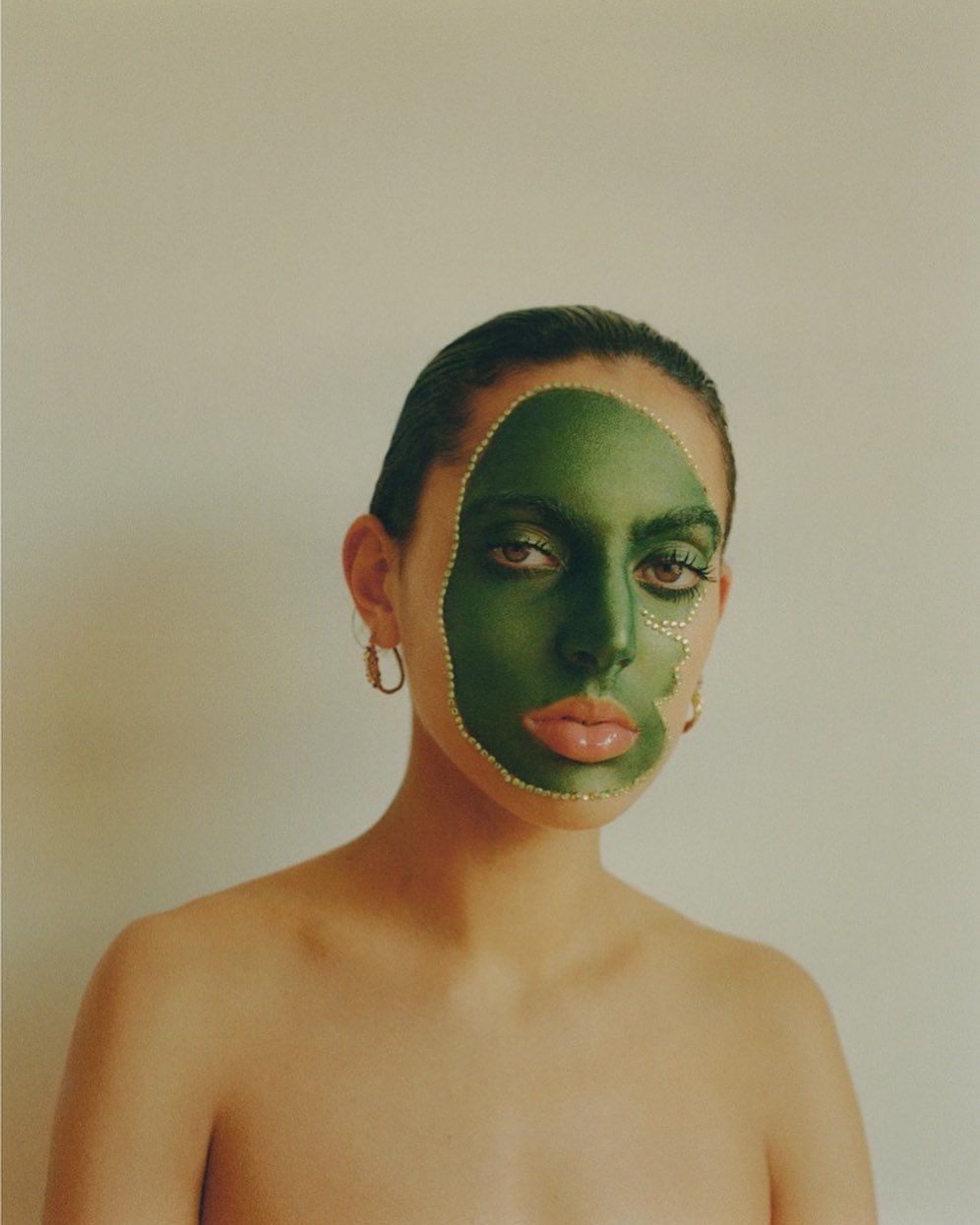 Portrait of a young woman with a green abstract makeup design outlined with pearls, accessorized with gold hoop earrings.