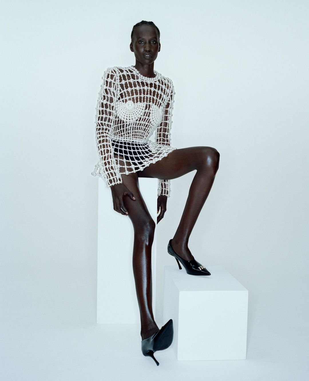 Model wearing a beaded mesh dress and black heels seated on white blocks against a plain background