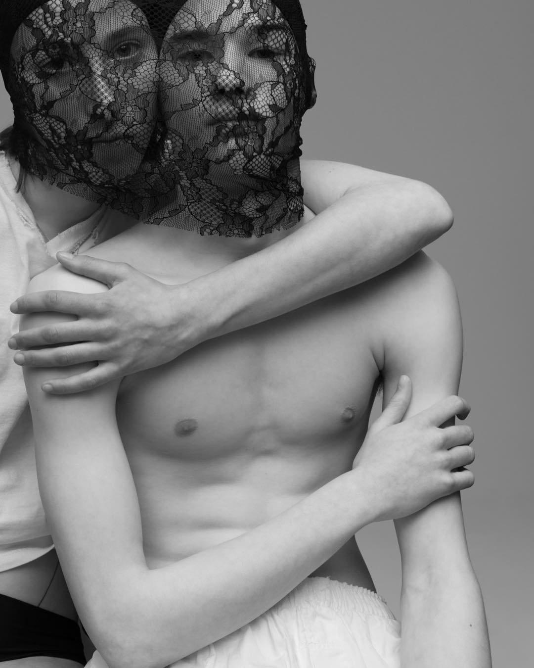 Two individuals with lace masks pose in a studio, their torsos bare, emphasizing texture and contrasting light and shadow in a monochrome portrait.