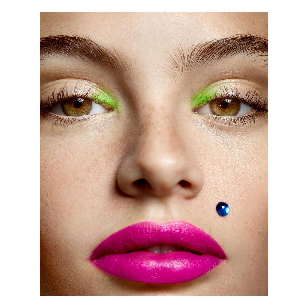 Extreme close-up of a fair-skinned woman with green neon eye makeup and bold pink lipstick with a blue gem on her cheek.
