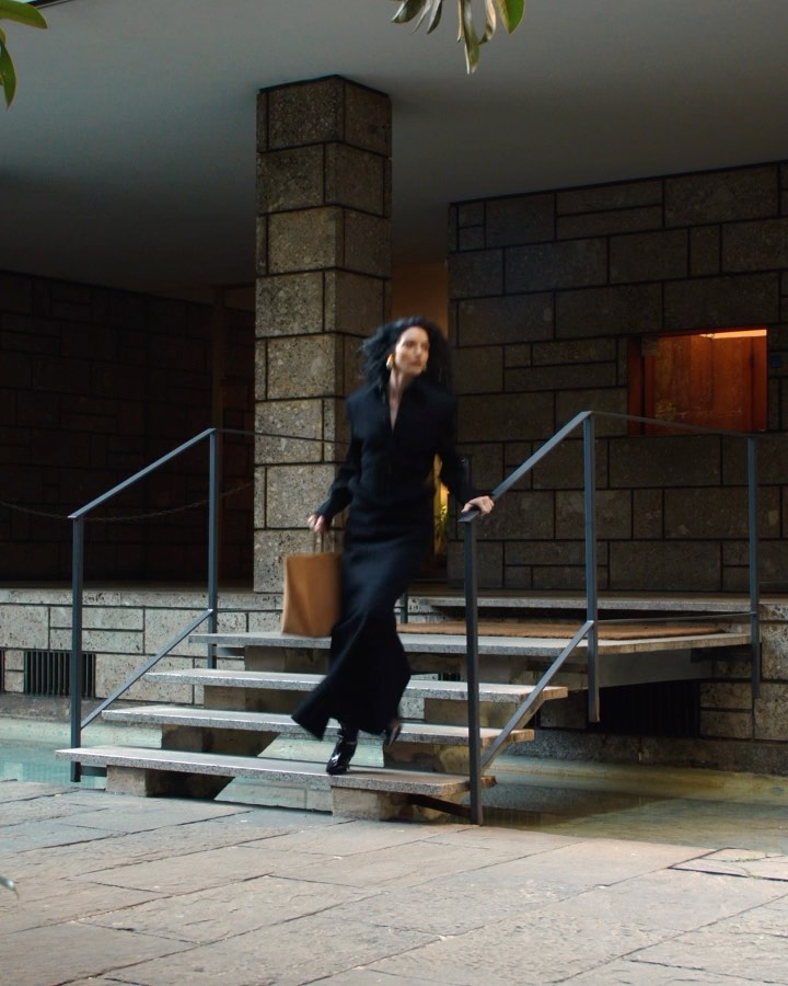 Elegant woman in a black dress descends stone steps of a modern building holding a tan tote bag, captured in a medium shot.
