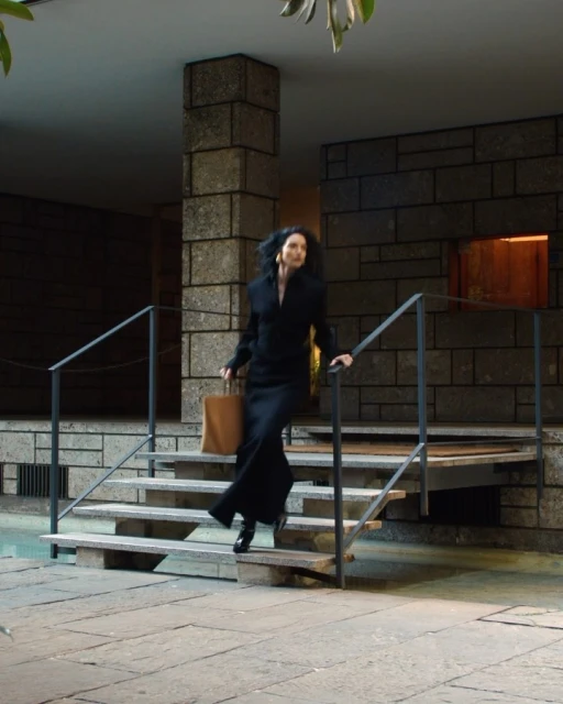 Elegant woman in a black dress descends stone steps of a modern building holding a tan tote bag, captured in a medium shot.
