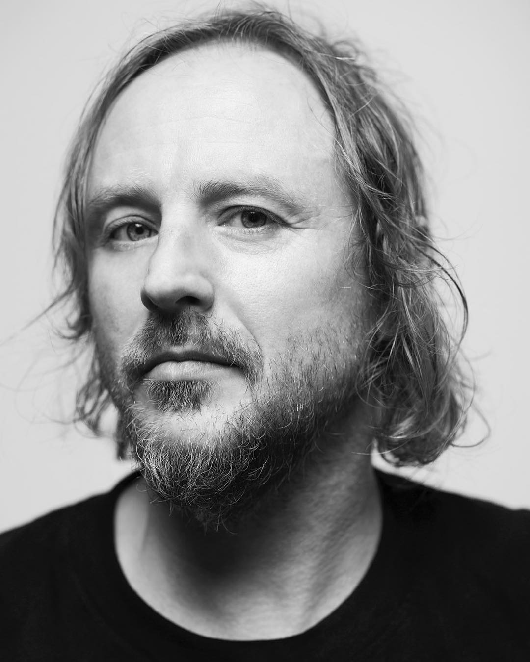 Black and white portrait of an adult male with medium-length hair and a beard, wearing a dark shirt, captured in a close-up studio shot.