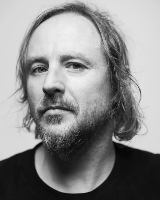Black and white portrait of an adult male with medium-length hair and a beard, wearing a dark shirt, captured in a close-up studio shot.