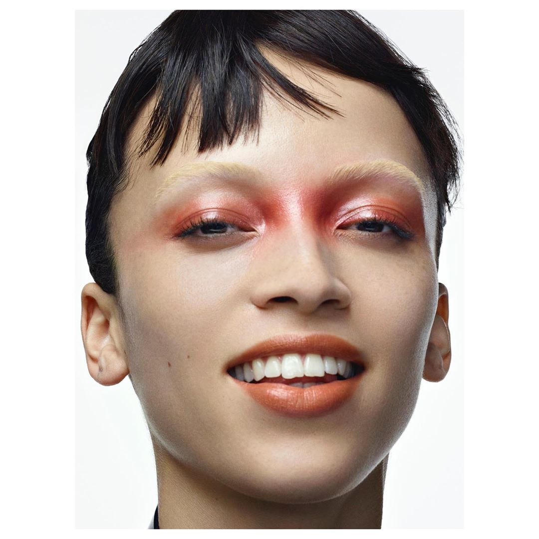 Close-up studio shot of a smiling adult female with a fair complexion and bold red and orange makeup around her eyes and mouth.