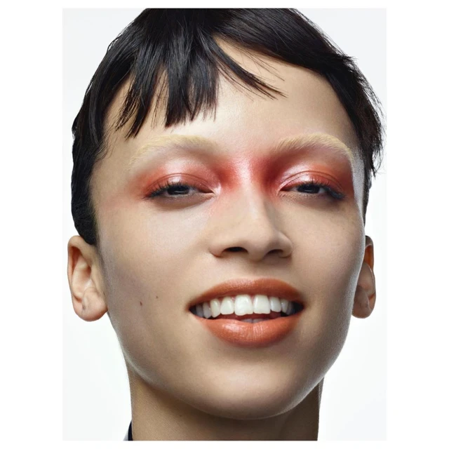 Close-up studio shot of a smiling adult female with a fair complexion and bold red and orange makeup around her eyes and mouth.