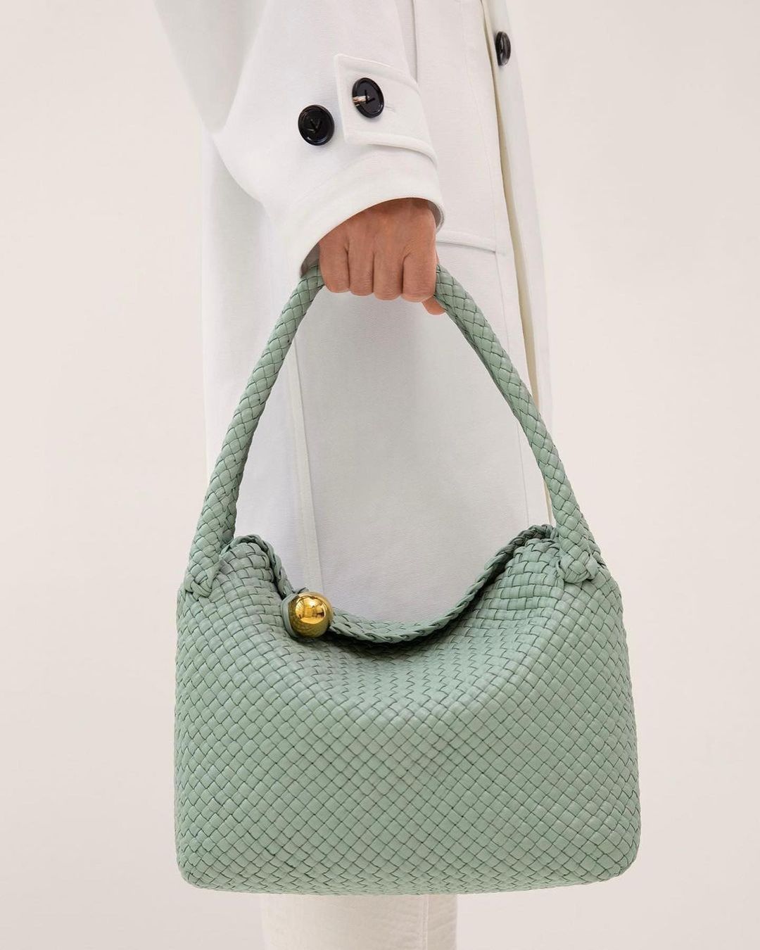Close-up of a light green woven leather handbag being held by an adult female wearing a white coat against an off-white background.