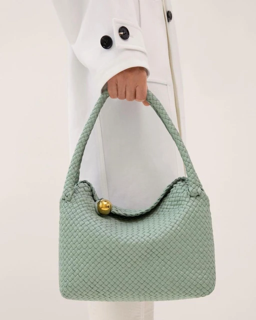 Close-up of a light green woven leather handbag being held by an adult female wearing a white coat against an off-white background.