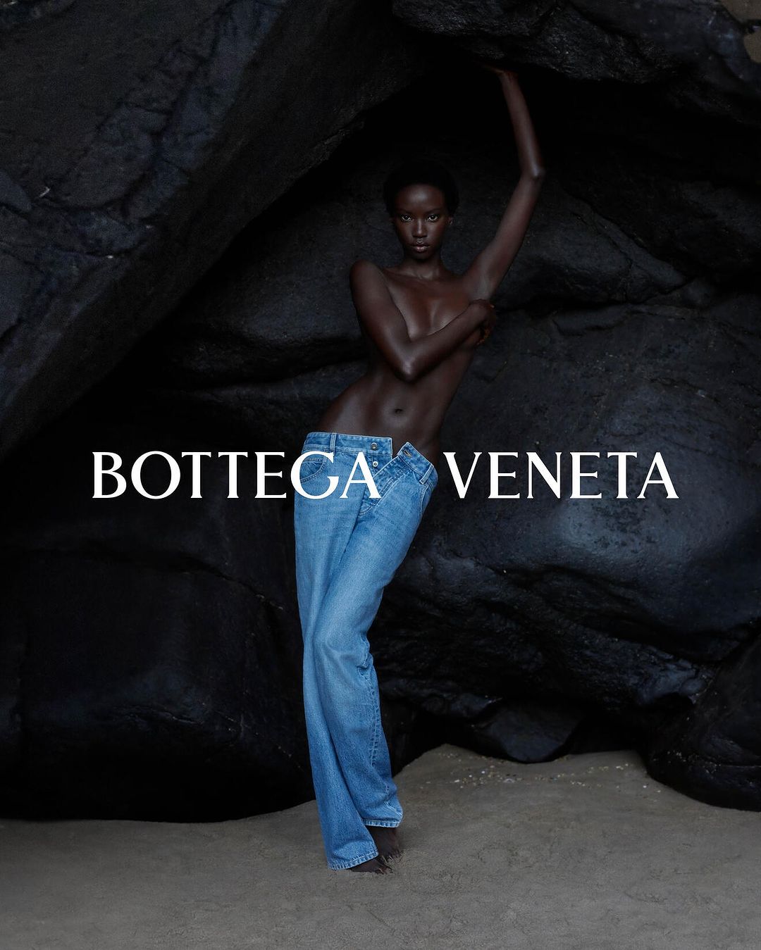 A woman posing in jeans against a rock formation, showcasing fashion, beauty, and editorial style with Bottega Veneta branding.