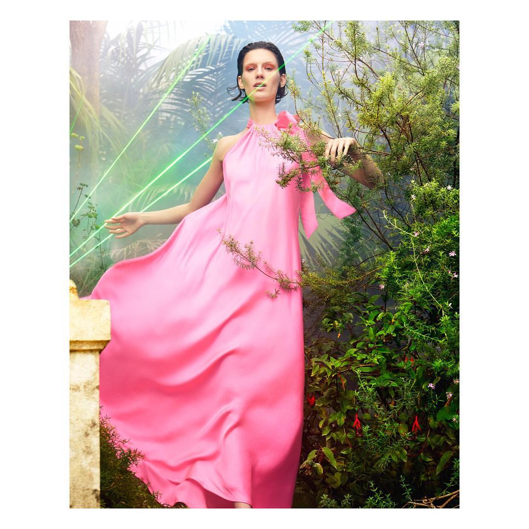 A woman in a pink gown poses in a garden, surrounded by greenery and with soft light filtering through the foliage.
