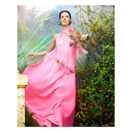 A woman in a pink gown poses in a garden, surrounded by greenery and with soft light filtering through the foliage.
