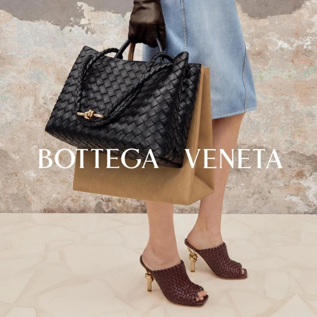 Fashionable woman showcases a black Bottega Veneta woven tote bag and matching heeled mules against a neutral marble backdrop.