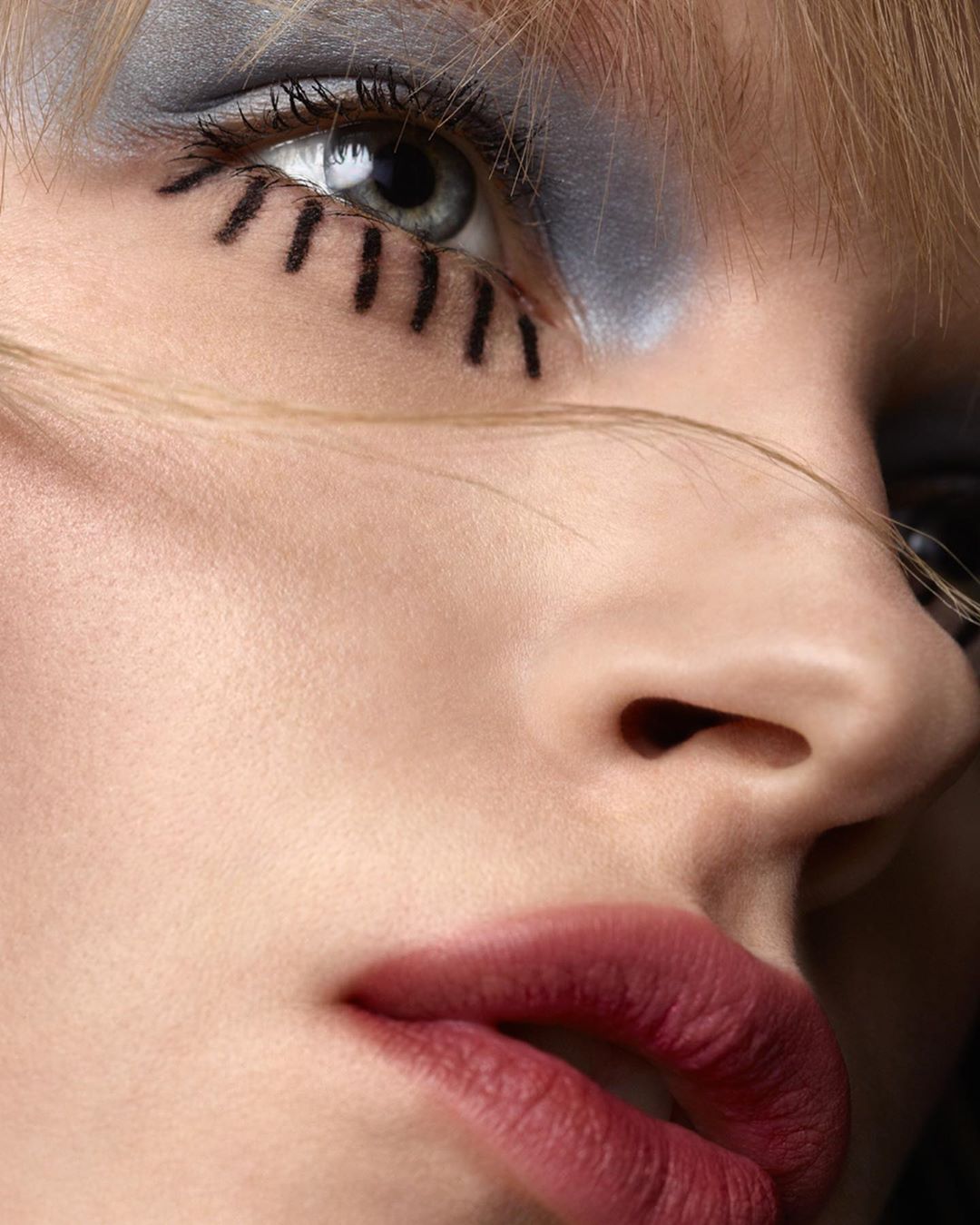 Close-up of a woman's face, showcasing detailed eye makeup with graphic black liner, grey eyeshadow, and bold dark red lipstick.