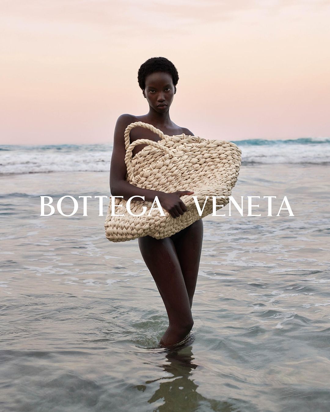 Fashion shot of an African woman holding a woven bag while standing in ocean water with a pink and orange sunset background.