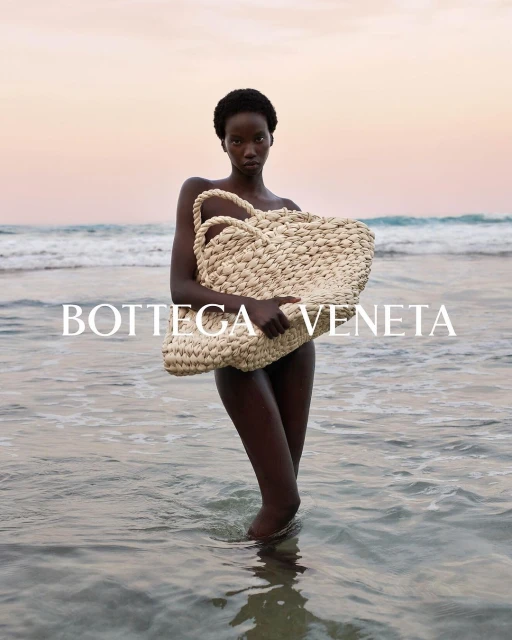 Fashion shot of an African woman holding a woven bag while standing in ocean water with a pink and orange sunset background.