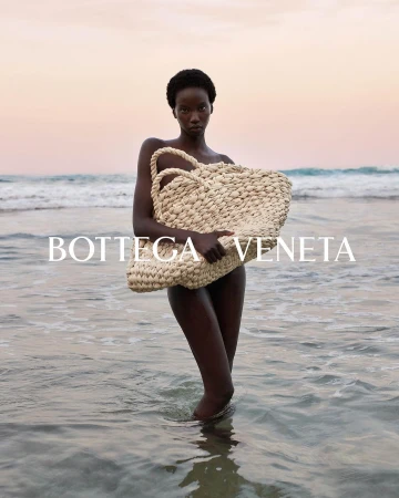Fashion shot of an African woman holding a woven bag while standing in ocean water with a pink and orange sunset background.