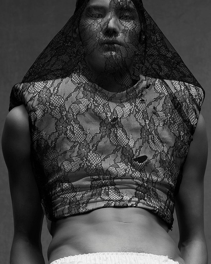 Monochrome portrait of a person with lace fabric draped over their head and body, posing in front of a gray background.