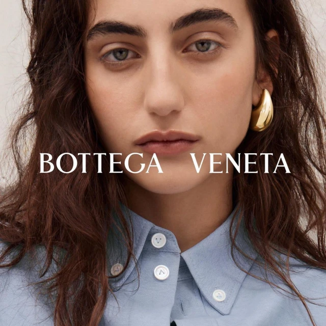 Close-up of a woman with auburn hair wearing a light blue button-down shirt and gold hoop earrings, featuring the Bottega Veneta logo.
