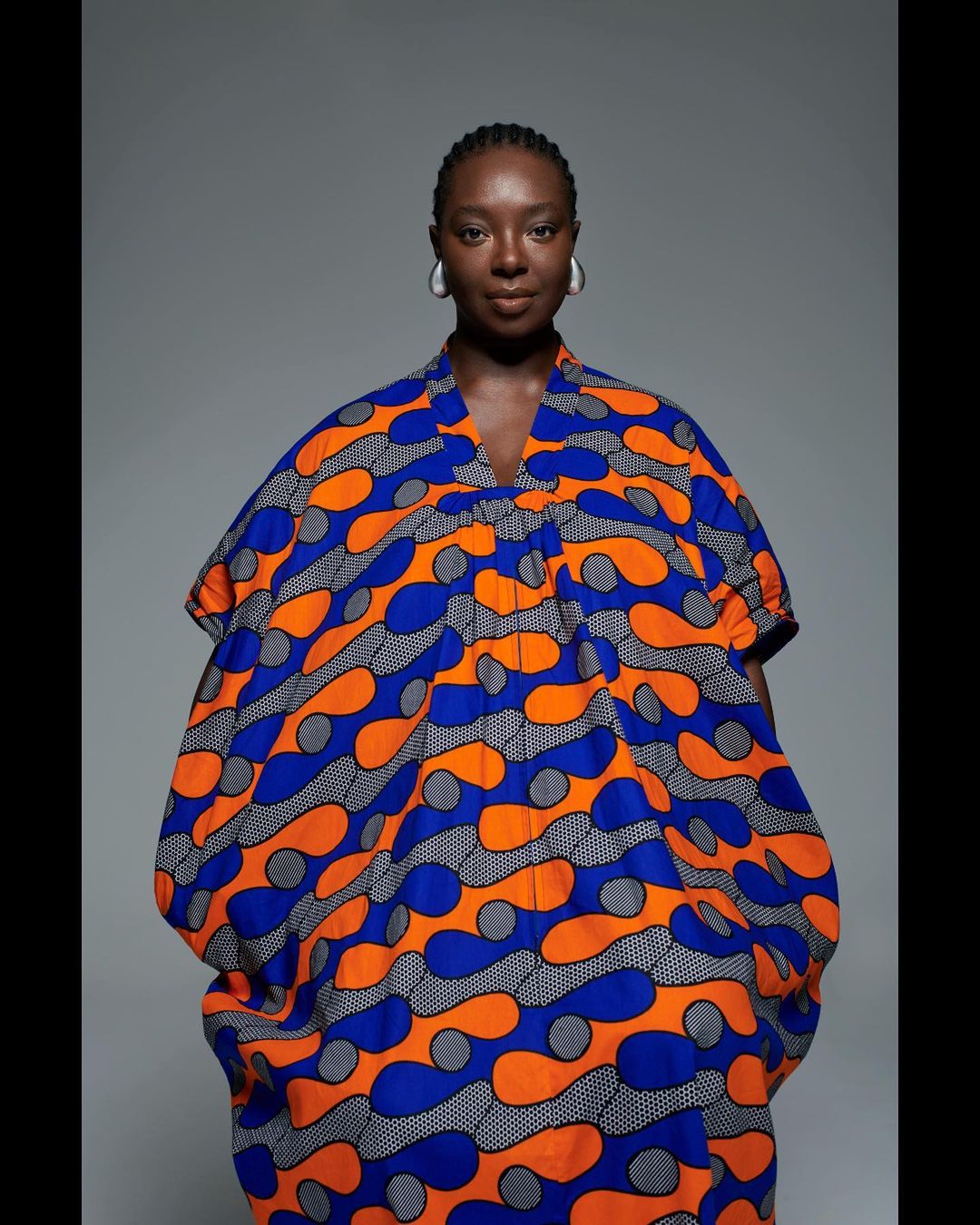 A solo adult female presents in a colorful patterned, relaxed-fit dress against a gray studio backdrop, showcasing beauty and fashion.