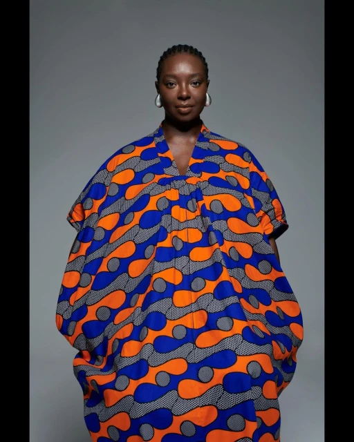 A solo adult female presents in a colorful patterned, relaxed-fit dress against a gray studio backdrop, showcasing beauty and fashion.